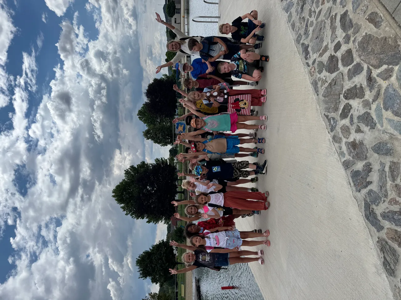 Eine Gruppe von Kindern und Erwachsenen, die zusammenstehen, lächeln und für ein Foto auf einem Pfad am See mit bewölktem Himmel posieren.