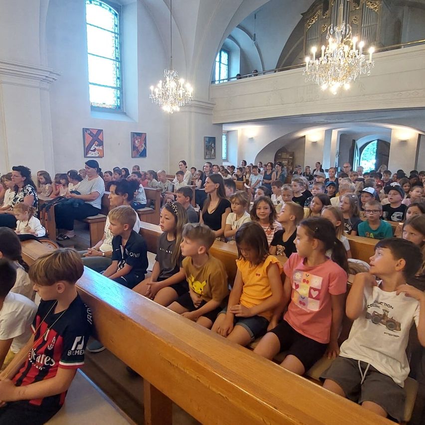 Eine große Gruppe von Kindern und Erwachsenen sitzt in einer Kirche, mit Kronleuchtern an der Decke. Die Kinder sind in verschiedenen Sommerkleidungen gekleidet, und einige tragen Halsketten.