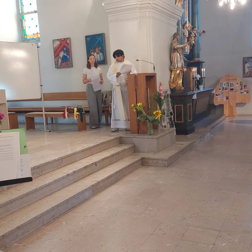 Ein Priester liest von einem Podium in einer Kirche, eine Frau steht neben ihm und hält Papiere. Dahinter steht eine Statue eines Heiligen, und nebenan befindet sich eine Holzstruktur mit Bildern.