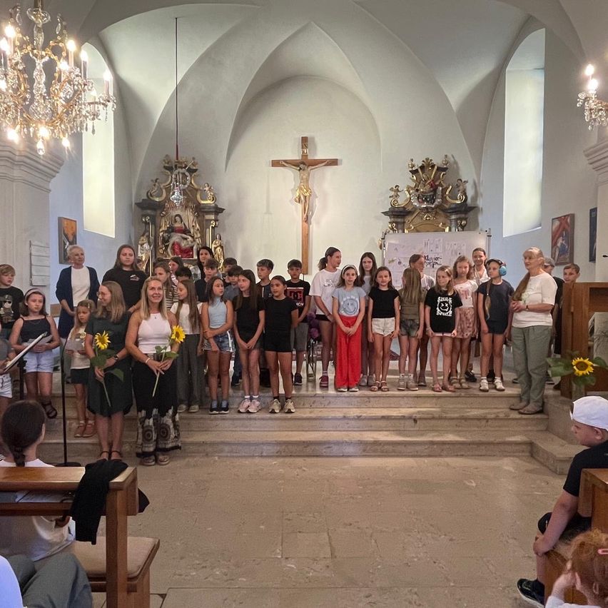 Eine Gruppe von Kindern und Erwachsenen steht in einer Kirche, hält Blumen und schaut zum Altar mit einem Kreuz.
