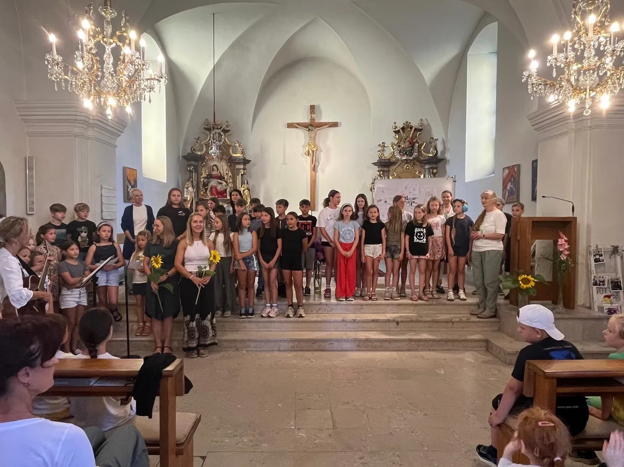 Eine Gruppe von Kindern und Erwachsenen steht in einer Kirche, hält Blumen und schaut zum Altar mit einem Kreuz.