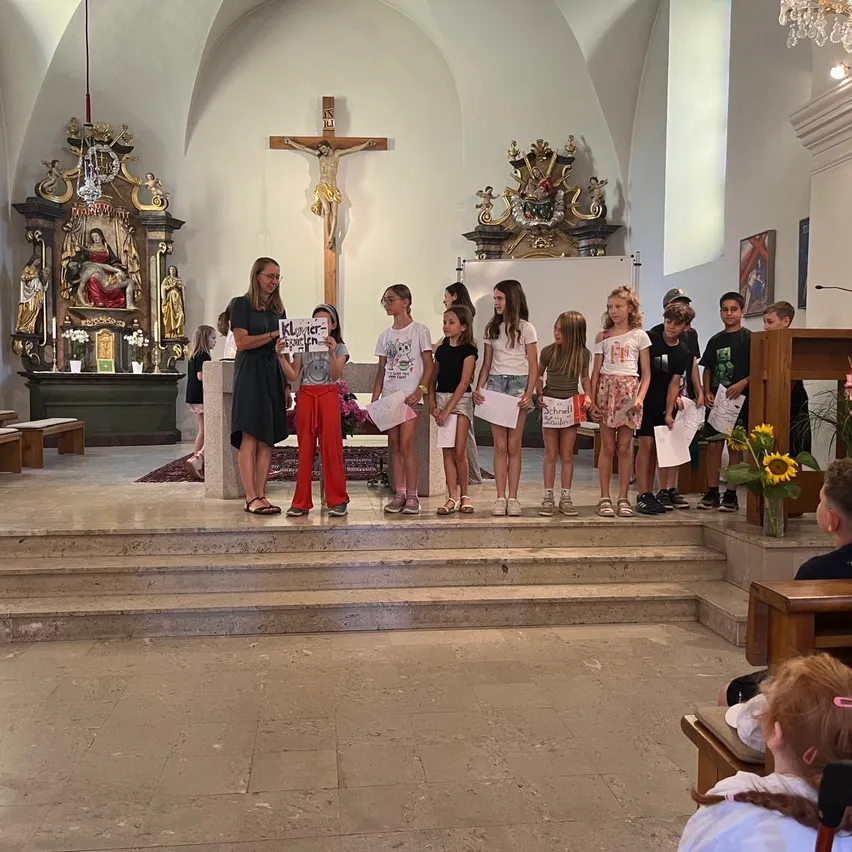 Eine Gruppe von Kindern und einer Frau steht auf den Stufen einer Kirche, hält Papiere und Schilder. Die Frau trägt eine Brille und ein schwarzes Kleid. Dahinter sind ein Kreuz und eine Statue zu sehen.