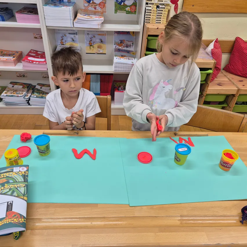 Zwei Kinder sitzen an einem Tisch im Klassenzimmer. Der Junge hält einen roten Buchstaben W und das Mädchen einen blauen Buchstaben W. Sie spielen mit Play-Doh und Buchstaben.