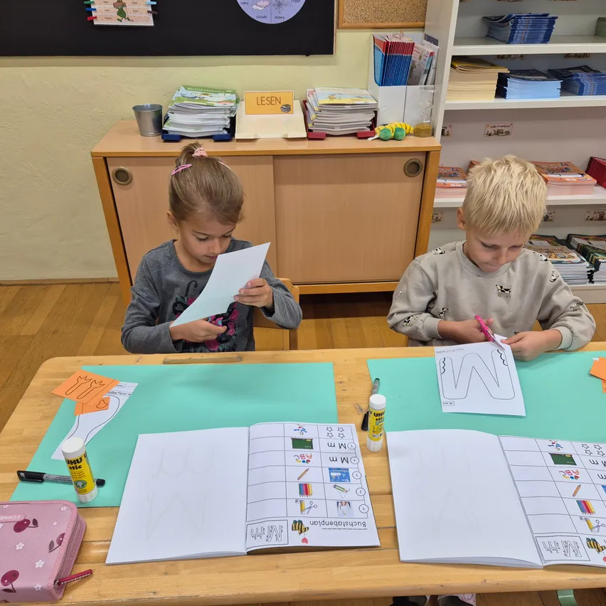 Zwei Kinder, ein Mädchen und ein Junge, sitzen an einem Tisch in einem Klassenzimmer. Das Mädchen hält ein Stück Papier in der Hand und schaut darauf, während der Junge einen Stift hält und auf einem Papier zeichnet. Hinter ihnen stehen Bücher in den Regalen und eine Pinnwand mit einem runden Diagramm an der Wand.