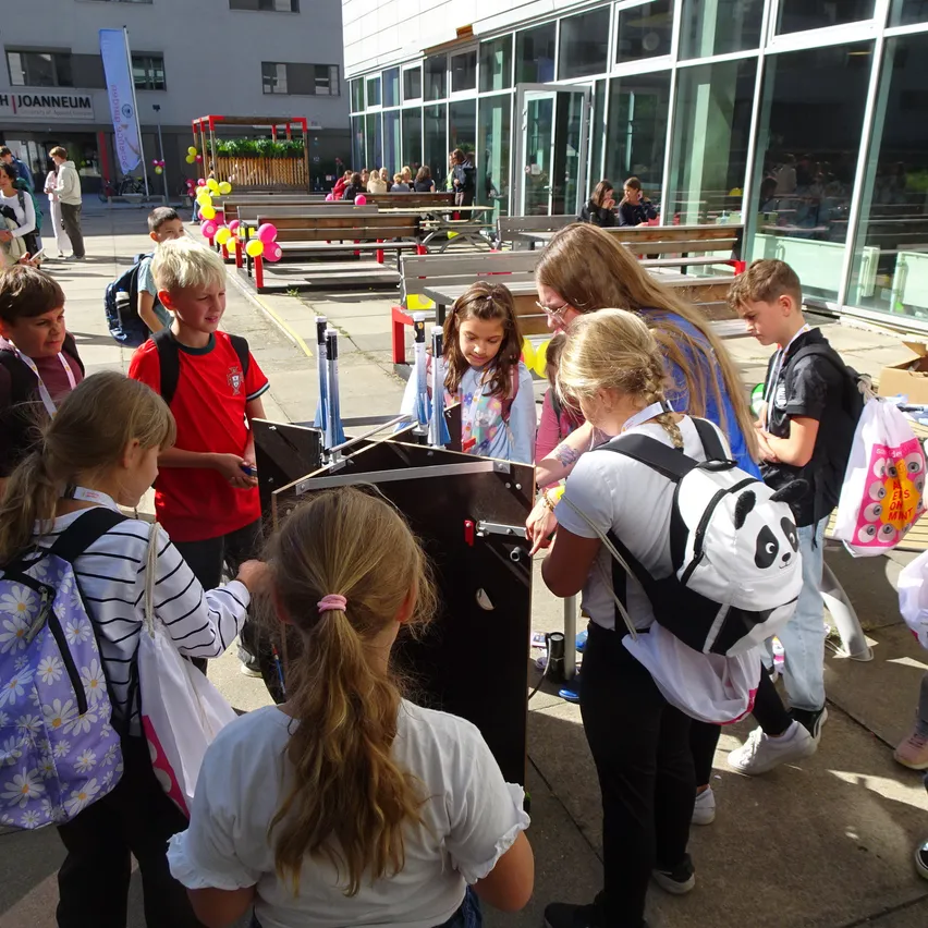 Eine Gruppe von Kindern versammelt sich um eine schwarze Box mit einem Panda-Design auf einem Rucksack. Sie befinden sich in einem Außenbereich, möglicherweise einer Schule.