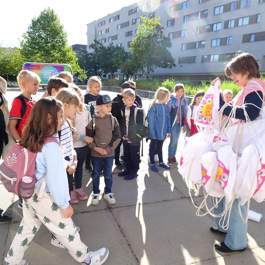Eine Gruppe von Kindern und Erwachsenen versammelt sich vor einem Gebäude. Die Kinder halten Taschen und scheinen auf etwas zu warten. Die Erwachsenen stehen hinter den Kindern.