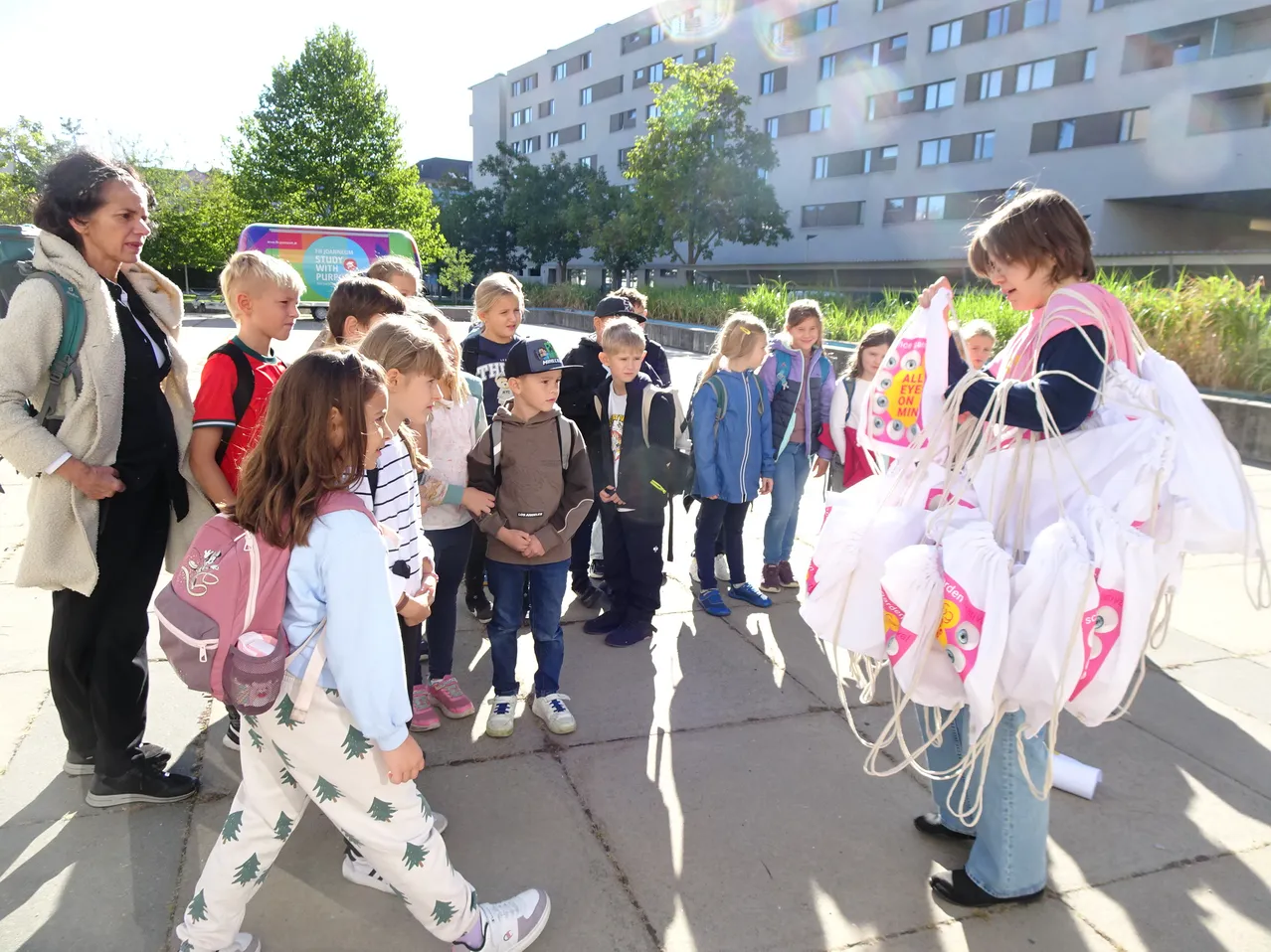 Eine Gruppe von Kindern und Erwachsenen versammelt sich vor einem Gebäude. Die Kinder halten Taschen und scheinen auf etwas zu warten. Die Erwachsenen stehen hinter den Kindern.