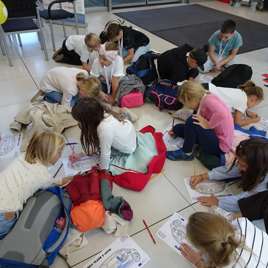 Eine Gruppe von Kindern sitzt auf dem Boden und zeichnet auf Papier. Einige tragen Rucksäcke und Schuhe.