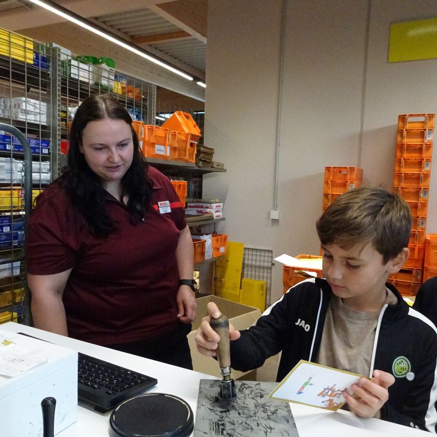 Eine Frau in einem weinroten Hemd steht hinter einem Jungen in einem schwarzen und weißen Jacke, der einen Stempel auf einem Stück Papier auf einem Tisch benutzt.
