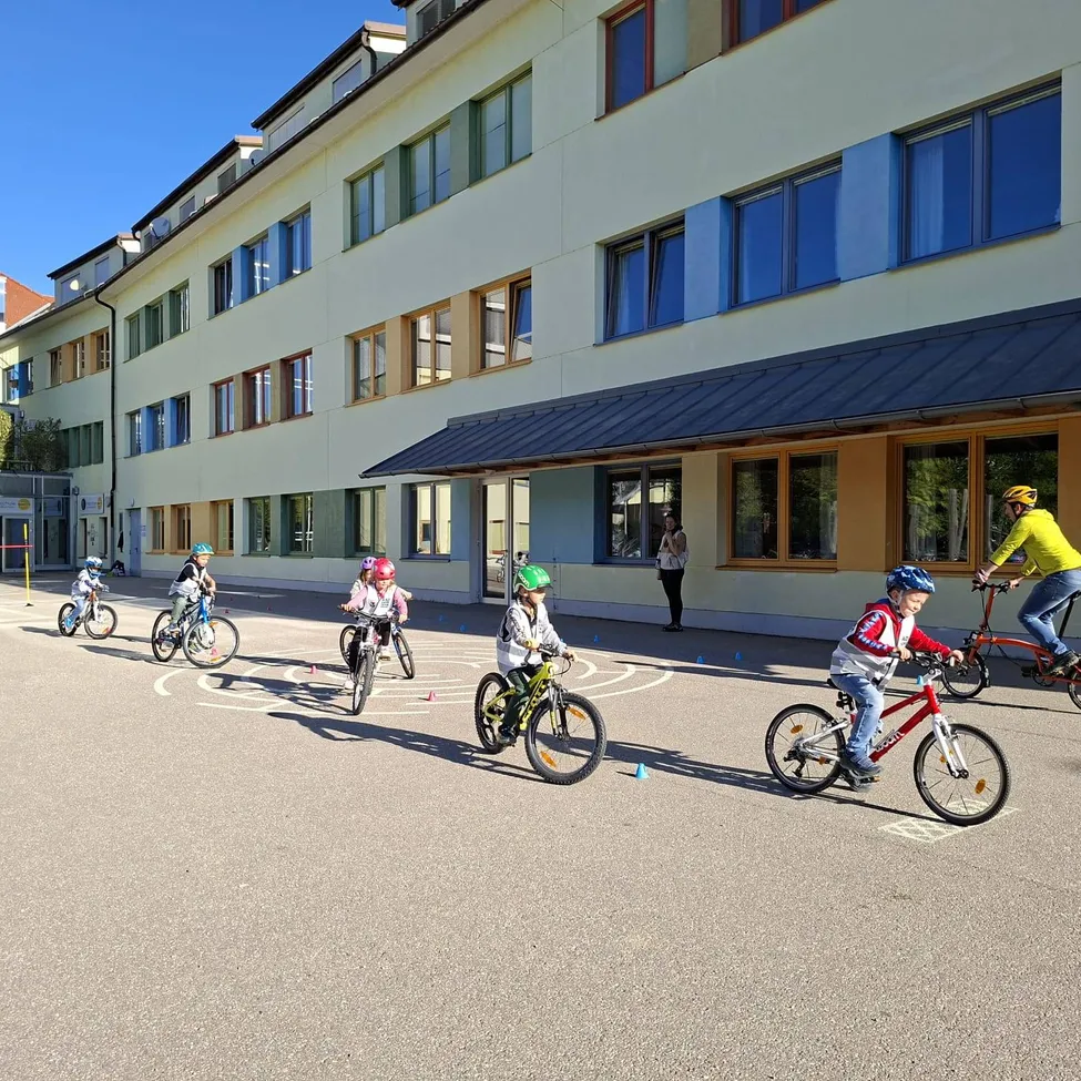 Eine Gruppe von Kindern fährt auf einem gepflasterten Bereich vor einem Gebäude im Kreis mit Fahrrädern.