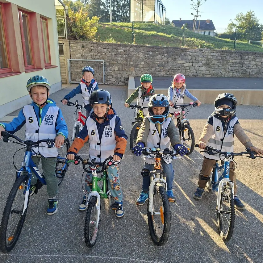 Eine Gruppe von Kindern, alle mit Helmen und Fahrradausrüstung, stehen in einer Reihe mit ihren Fahrrädern. Sie sind draußen auf einem Parkplatz, mit einem Gebäude und einer Steinmauer im Hintergrund.