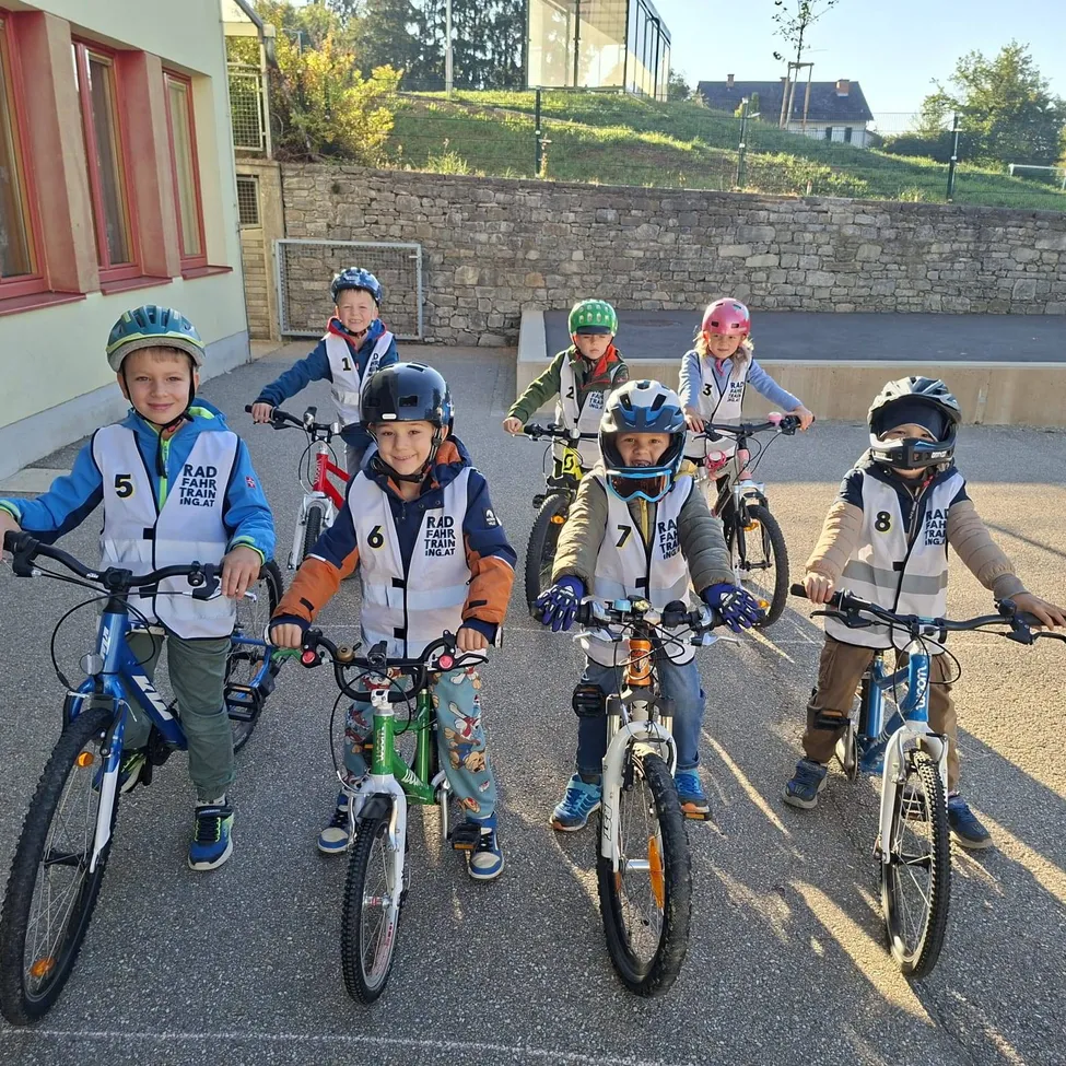 Eine Gruppe von Kindern, alle mit Helmen und Fahrradausrüstung, stehen in einer Reihe mit ihren Fahrrädern. Sie sind draußen auf einem Parkplatz, mit einem Gebäude und einer Steinmauer im Hintergrund.