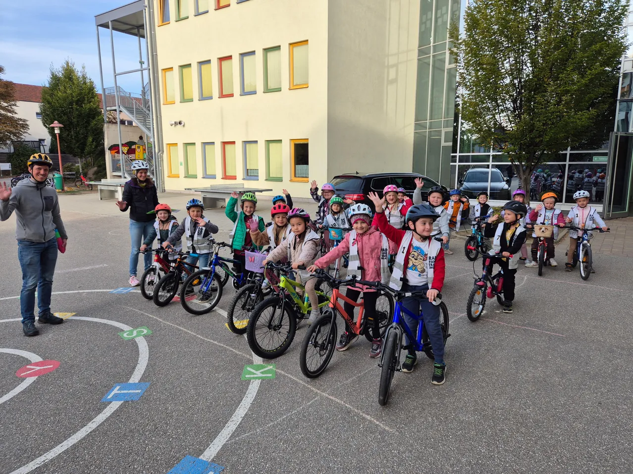 Eine Gruppe von Kindern mit Helmen fährt in einem Kreis auf Fahrrädern vor einem Gebäude.
