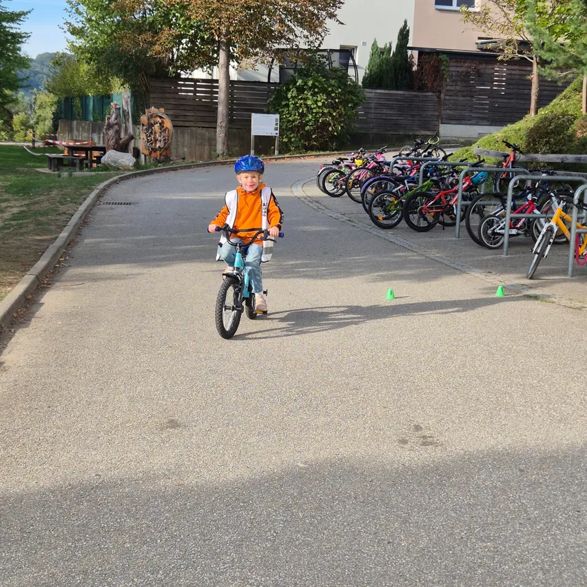 Ein junger Junge fährt auf einem asphaltierten Weg mit dem Fahrrad. Er trägt einen Helm und einen Rucksack. Am Straßenrand sind mehrere Fahrräder geparkt.