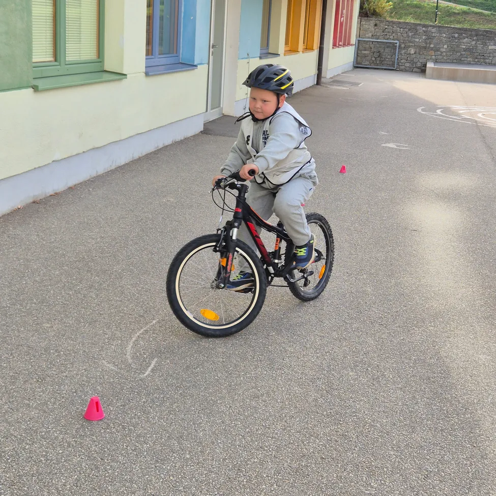 Ein junger Junge fährt auf einem gepflasterten Bereich ein Fahrrad. Er trägt einen Helm und Turnschuhe. Es gibt zwei rote Kegel auf dem Boden. Hinter ihm ist ein Gebäude mit gelben und blauen Fenstern und einem grauen Dach.