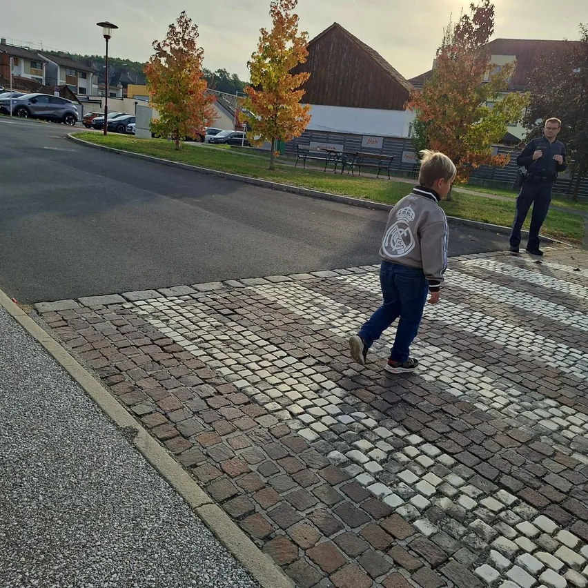 Ein junger Junge geht auf einem gepflasterten Gehweg und trägt eine Jacke mit einem weißen Logo. Hinter ihm steht ein Mann auf dem Bürgersteig.