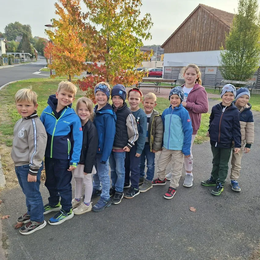 Eine Gruppe von Kindern, alle in Jacken gekleidet, steht in einer Reihe auf einer gepflasterten Straße. Hinter ihnen befinden sich Häuser, Bäume und eine Bank.
