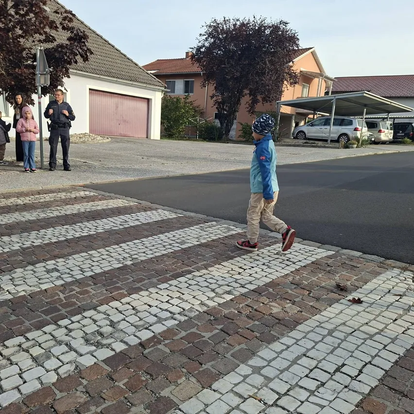 Ein kleiner Junge läuft über einen gepflasterten Zebrastreifen in einem Wohngebiet. Dahinter stehen eine Gruppe von Kindern und ein Polizist.