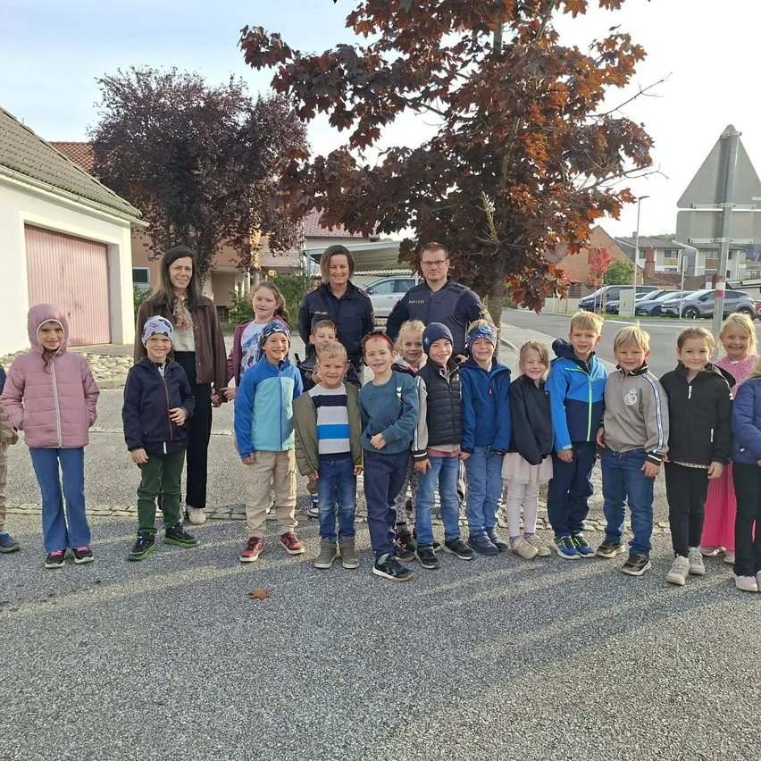 Eine Gruppe von Kindern und zwei Erwachsenen stehen in einer Reihe auf einer Straße. Die Kinder tragen Winterkleidung und die Erwachsenen Jacken. Im Hintergrund befinden sich Häuser und Bäume.