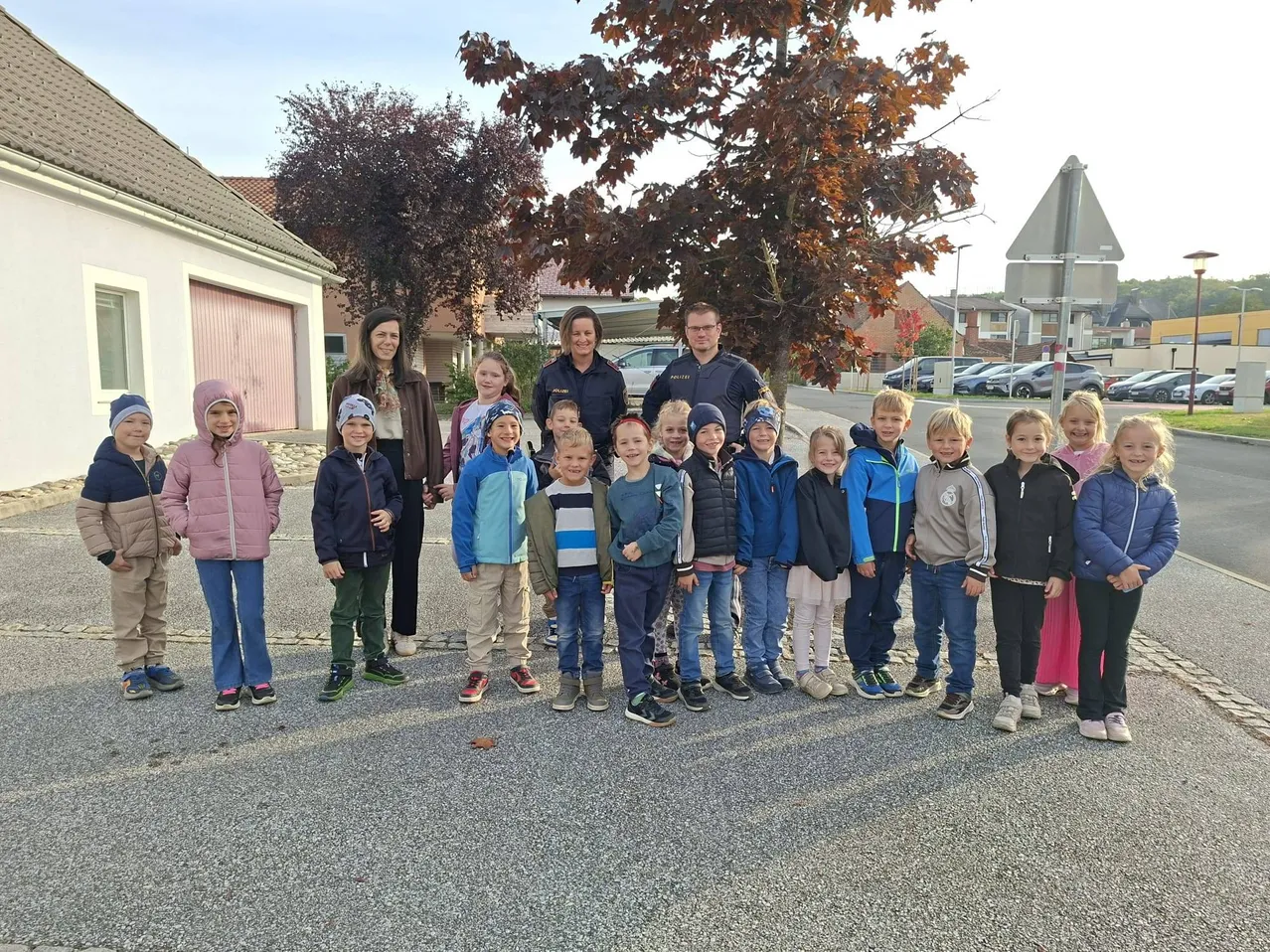Eine Gruppe von Kindern und zwei Erwachsenen stehen in einer Reihe auf einer Straße. Die Kinder tragen Winterkleidung und die Erwachsenen Jacken. Im Hintergrund befinden sich Häuser und Bäume.