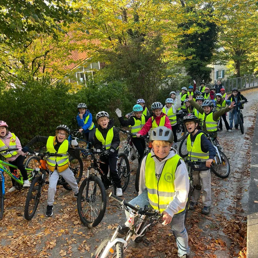 Eine Gruppe von Kindern, die neongelbe Westen und Helme tragen, fährt mit ihren Fahrrädern auf einem von Bäumen und Büschen umgebenen Pfad.