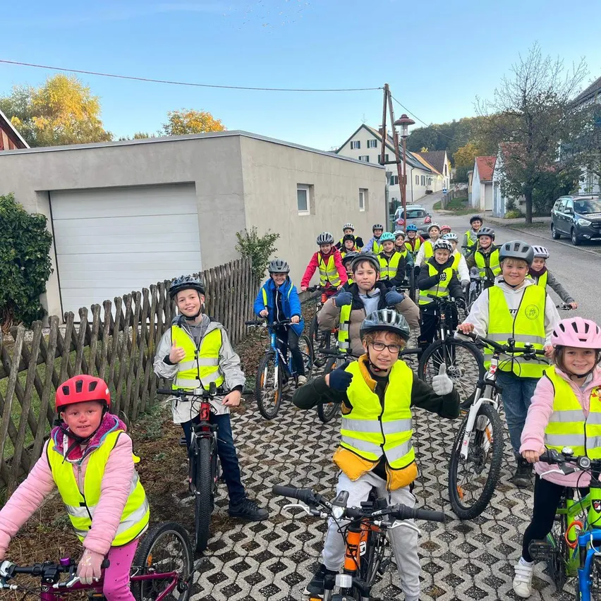 Eine Gruppe von Kindern, alle mit Sicherheitswesten und Helmen bekleidet, fahren auf einem gepflasterten Weg mit ihren Fahrrädern. Sie sind in Reihen aufgestellt, einige stehen und andere sitzen auf ihren Fahrrädern. Der Hintergrund zeigt ein Gebäude mit einer Garagentür und einem Baum.