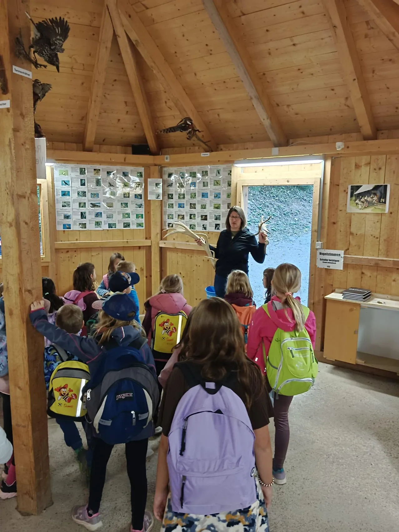 Eine Frau unterrichtet eine Gruppe von Kindern in einem hölzernen Raum. Die Kinder tragen Rucksäcke und hören der Frau zu, die ein Stück Holz in der Hand hält.