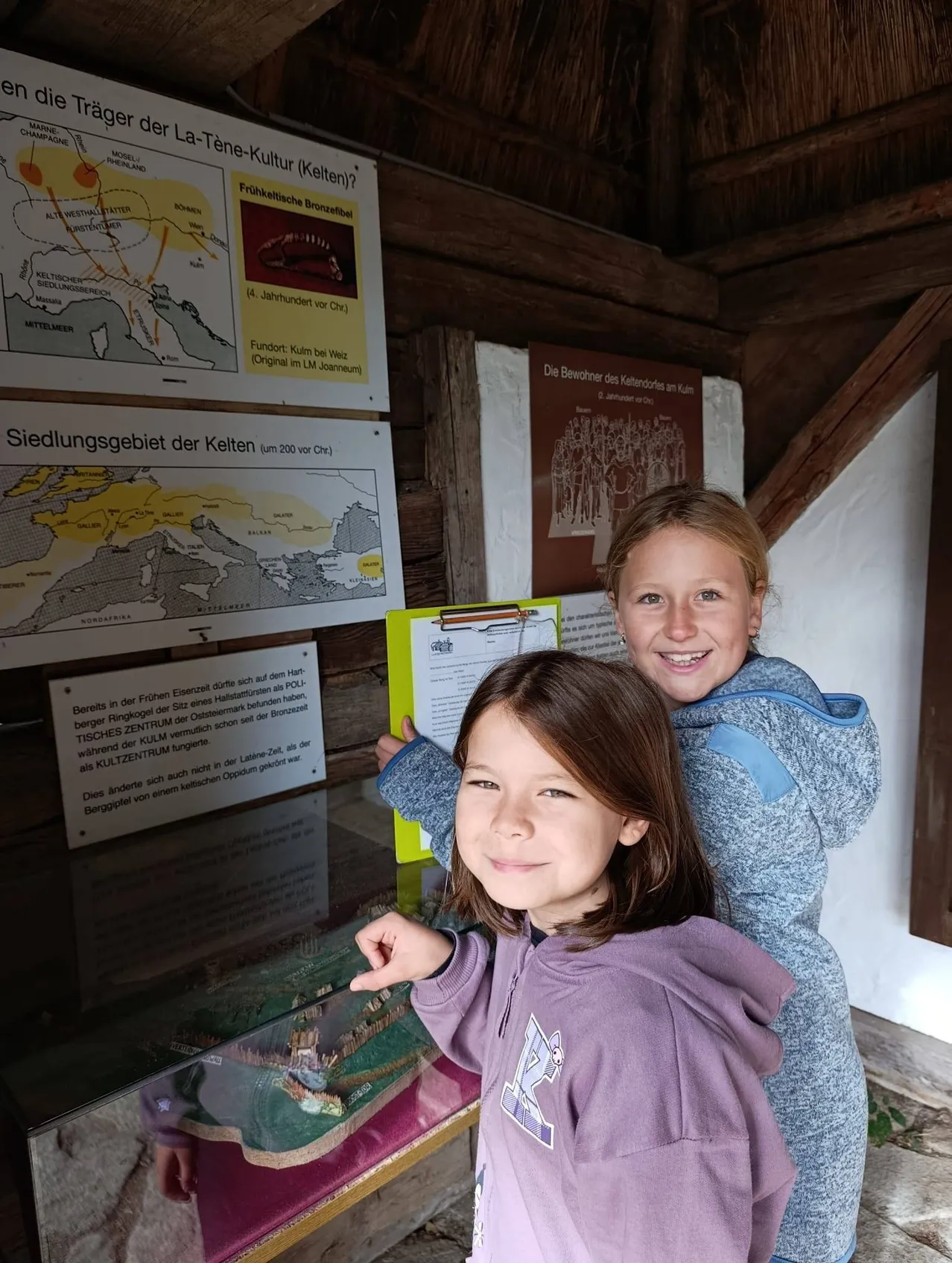 Zwei junge Mädchen stehen vor einer Vitrine mit einem Modell einer historischen Stätte. Die Vitrine befindet sich in einem kleinen Raum mit Holzwänden und einer Holzdecke.