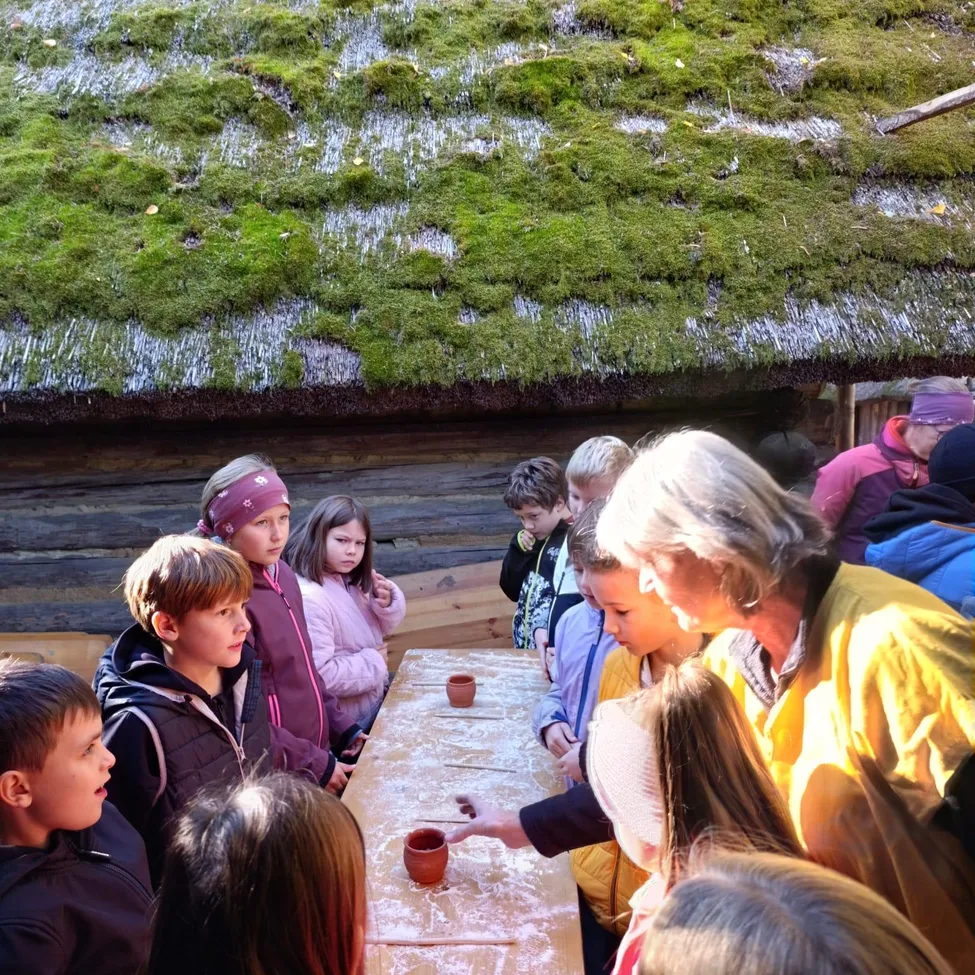 Eine Gruppe von Kindern und Erwachsenen versammelt sich um einen Tisch, möglicherweise in einem Museum oder einer Bildungseinrichtung. Der Tisch hat ein moosbedecktes Dach und eine Holzstruktur. Die Kinder schauen auf den Tisch, und einer von ihnen hält einen kleinen Becher.
