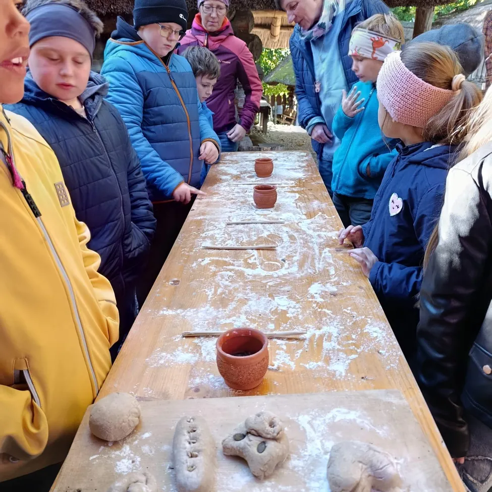 Eine Gruppe von Menschen ist um einen Holztisch versammelt und beschäftigt sich mit einer Töpferaktivität. Der Tisch ist mit Tonskulpturen bedeckt und die Teilnehmer tragen Winterkleidung.