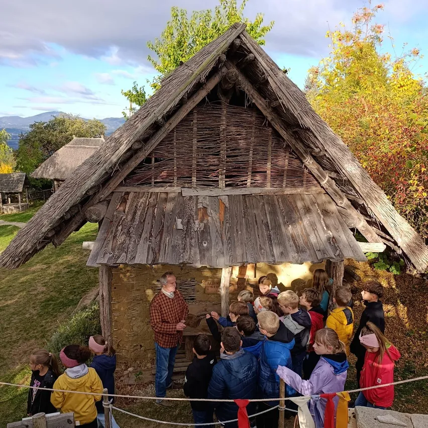 Eine Gruppe von Kindern und ein erwachsener Mann versammeln sich um ein kleines, altes, hölzerne Haus mit einem Strohdach. Das Haus ist von Bäumen und Gras umgeben.