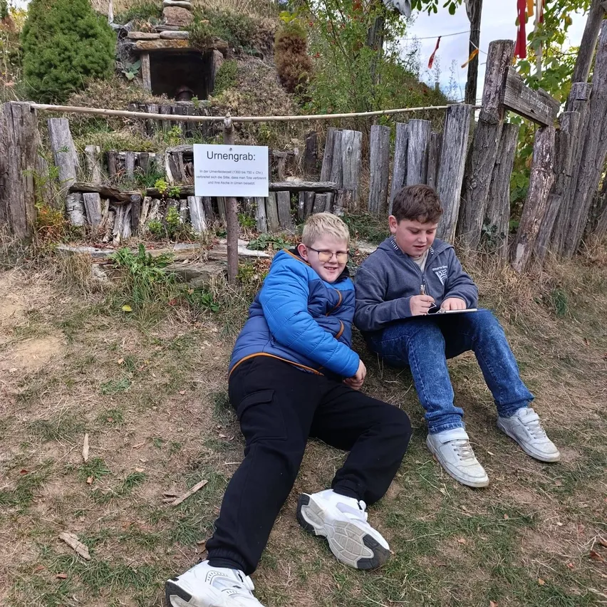 Zwei Jungen sitzen auf dem Gras vor einem Holzzaun. Ein Junge trägt eine Brille und eine blaue Jacke, während der andere Junge eine graue Jacke und Jeans trägt. Sie schauen beide lächelnd auf ein Tablet. Hinter ihnen steht ein Schild mit der Aufschrift 'Urmengengrab'.
