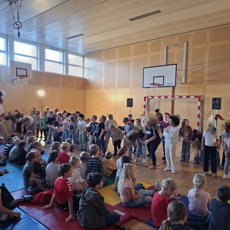 Eine Gruppe von Kindern sitzt auf dem Boden in einer Turnhalle und schaut einem Mann beim Gitarrespielen zu.