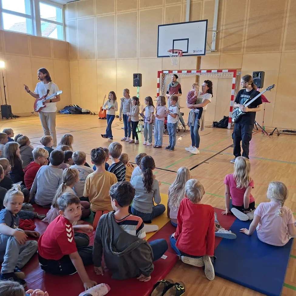 Eine Gruppe von Kindern und Erwachsenen versammelt sich in einer Turnhalle. Zwei Erwachsene stehen vor einer Gruppe von Kindern und halten Gitarren. Die Kinder sitzen auf Yogamatten und beobachten die Erwachsenen.