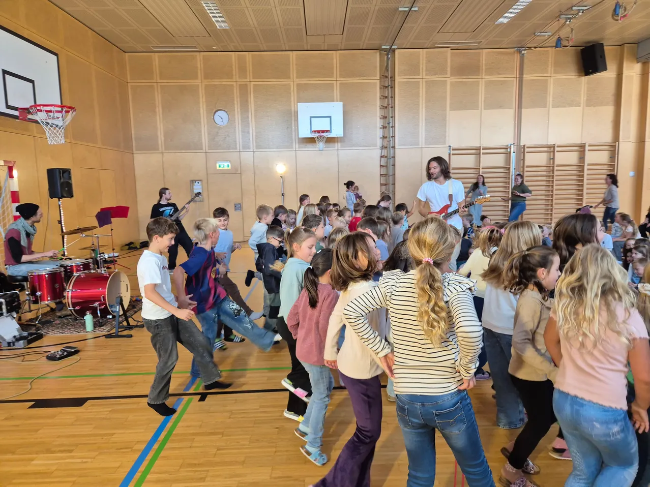 Eine Gruppe von Kindern und Erwachsenen tanzt in einer Turnhalle. Ein Mann spielt Gitarre und ein anderer Mann Schlagzeug.