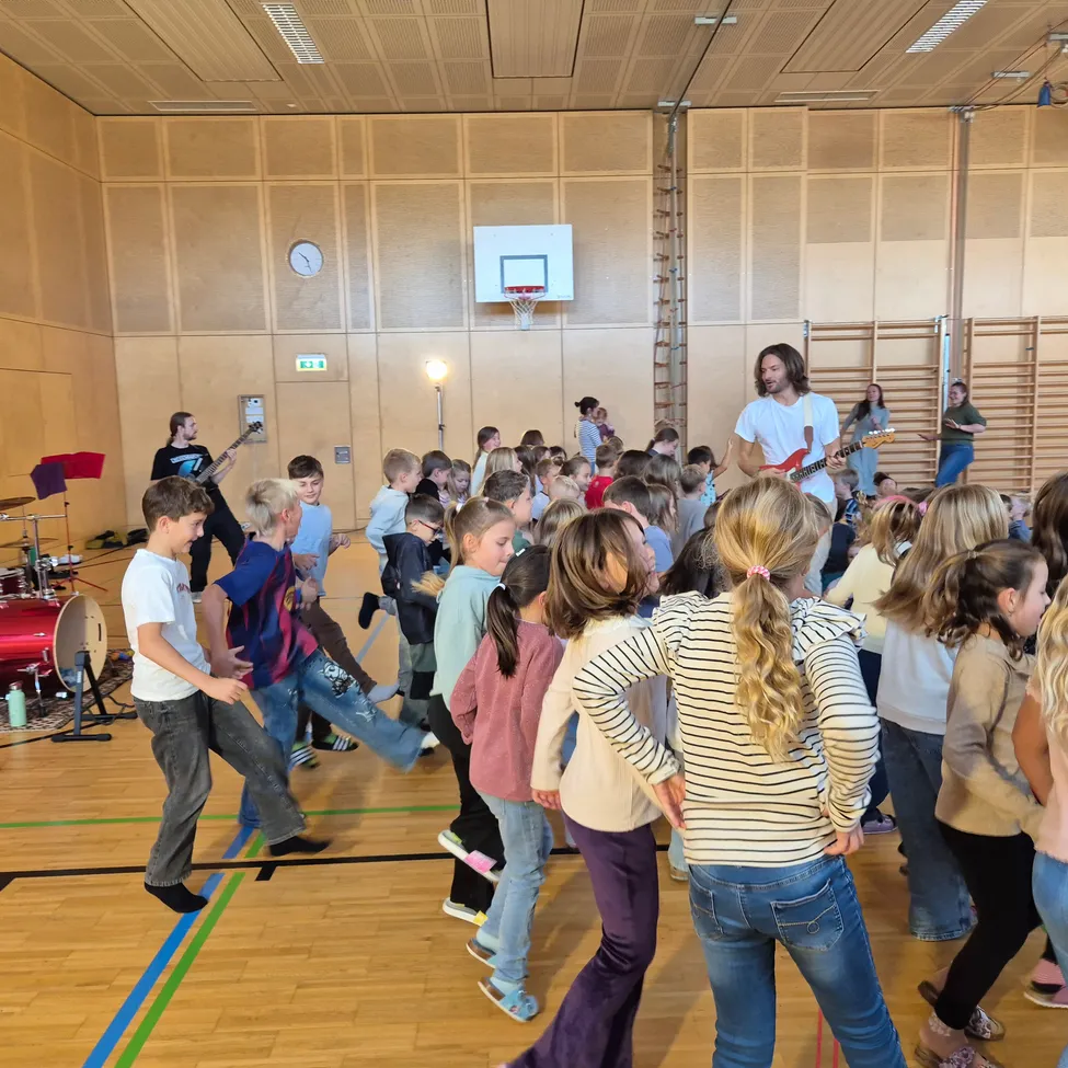 Eine Gruppe von Kindern und Erwachsenen tanzt in einer Turnhalle. Ein Mann spielt Gitarre und ein anderer Mann Schlagzeug.