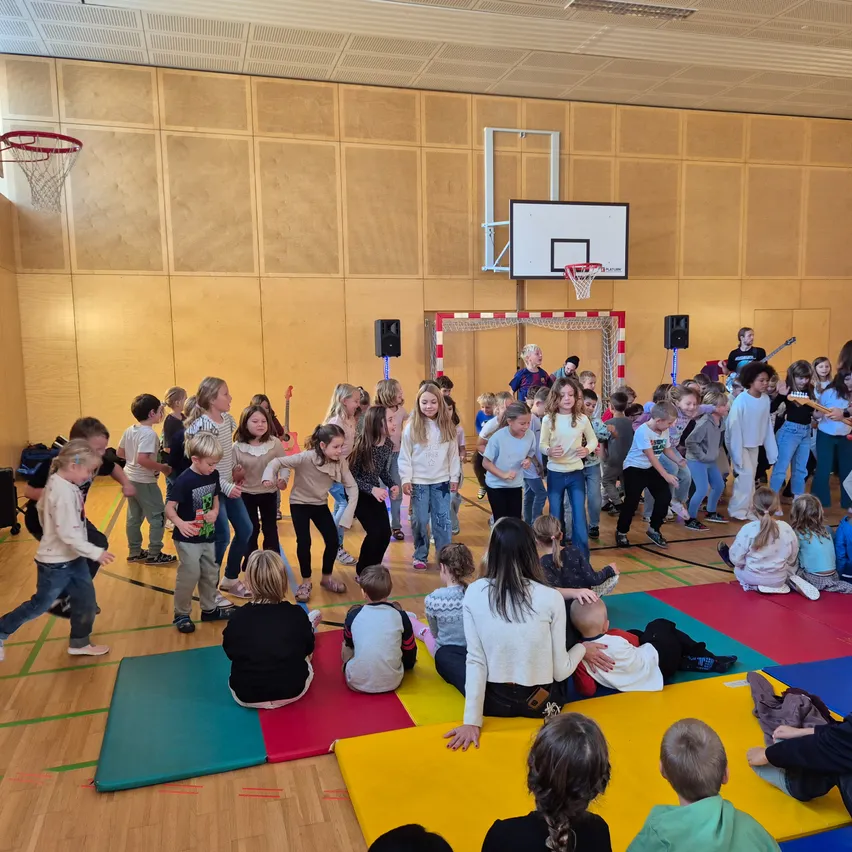 Eine Gruppe von Kindern versammelt sich in einer Turnhalle, einige sitzen auf bunten Matten, während andere stehen und tanzen. Einige Erwachsene spielen Gitarre und singen.