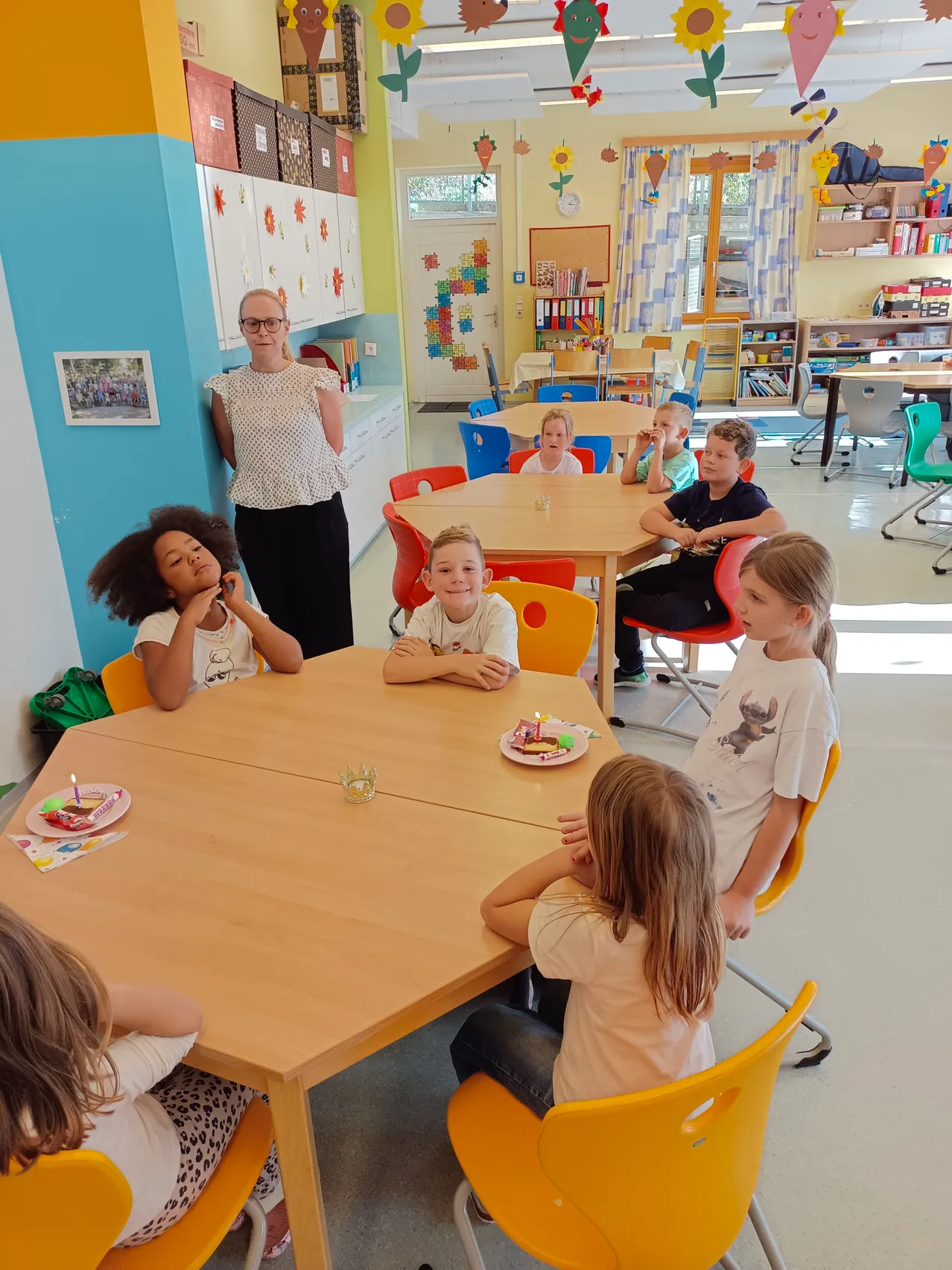 Eine Gruppe von Kindern sitzt um einen Tisch in einem Klassenzimmer, mit einer Frau, die hinter ihnen steht. Der Tisch hat Teller mit Geburtstagskuchen und Kerzen.