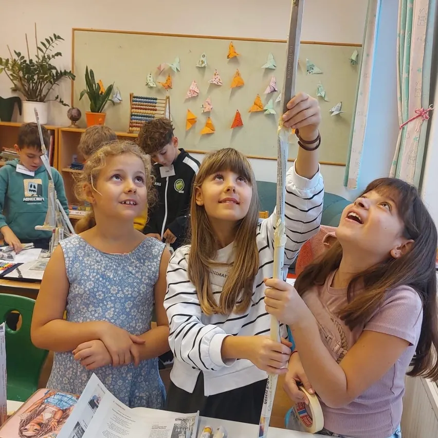 Vier Mädchen in einem Klassenzimmer, eines hält einen langen Stock, während die anderen interessiert zusehen.