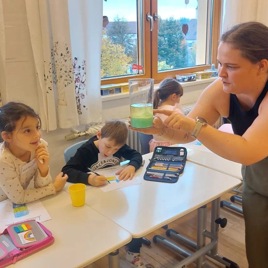 Eine Frau steht neben einem Schreibtisch und hält ein Glas Wasser in der Hand. Sie schaut ein junges Mädchen und einen Jungen an, die am Schreibtisch sitzen. Das Mädchen hält einen gelben Becher und der Junge einen Bleistift. Im Hintergrund sitzen andere Kinder an anderen Schreibtischen.