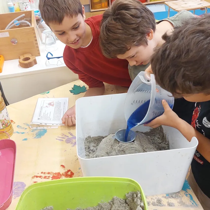 Drei junge Jungen sind in einer Sandkunstaktivität beschäftigt. Ein Junge gießt blaue Flüssigkeit in einen mit Sand gefüllten Behälter.
