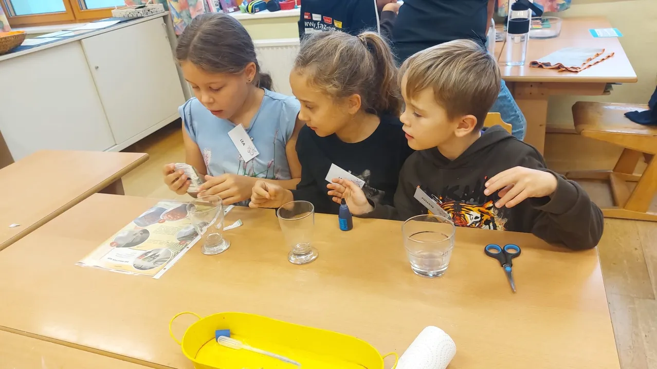 Drei Kinder sitzen an einem Tisch und sind in ein wissenschaftliches Experiment vertieft. Das Mädchen auf der linken Seite hält ein kleines Objekt, während das Mädchen in der Mitte ein Stück Papier in der Hand hat. Der Junge auf der rechten Seite hält eine kleine Flasche.