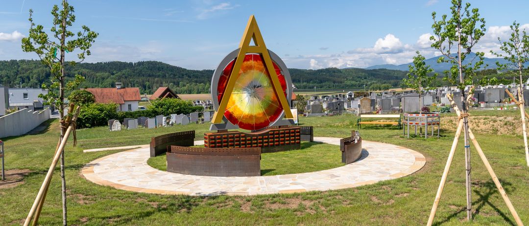Eine farbenfrohe, kreisförmige Skulptur auf einem Friedhof. Eine gelbe dreieckige Form oben. Umgeben von Bänken. Berge und Bäume im Hintergrund.