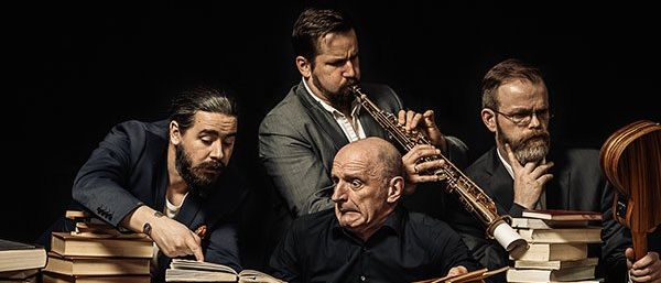 Vier Männer posieren in einem Studio, umgeben von einem Stapel Bücher, jeder hält ein Musikinstrument.