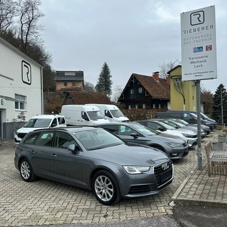 Ein grauer Audi-Wagen ist auf einem Parkplatz geparkt, mit einem Schild, das Rieberer Autohandel KFZ Technik anzeigt. Dahinter stehen mehrere andere Autos, Lieferwagen und eine Holzbank. In der Ferne sind Häuser und Bäume zu sehen.