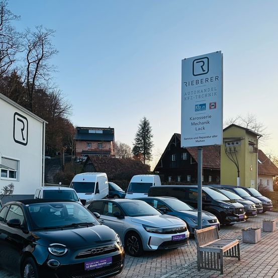 Ein Autohaus mit einem Schild und mehreren Autos und Lieferwagen davor. Eine Bank steht in der Nähe des Schildes. Der Himmel ist klar und blau.