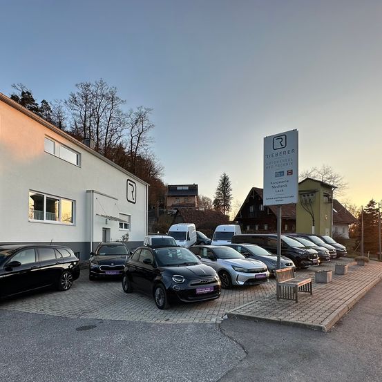 Mehrere Autos sind vor einem Gebäude geparkt, das mit 'Rieberer Autohandel Kfz-Technik' beschriftet ist. Im Hintergrund sind Bäume und Häuser zu sehen.