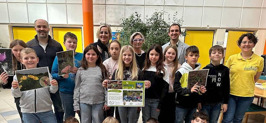Eine Gruppe von Kindern und Erwachsenen steht in einem Klassenzimmer und posiert für ein Foto. Die Kinder halten ein Zertifikat und ein Foto. Die Erwachsenen lächeln und posieren für ein Foto.
