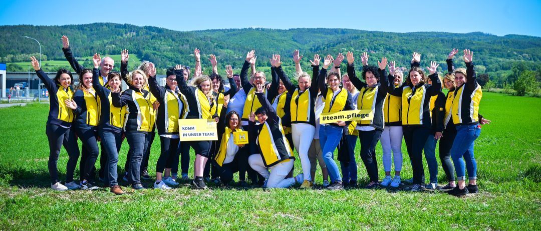Eine Gruppe von Menschen in gelben und schwarzen Jacken steht auf einem Feld, einige heben ihre Hände. Im Hintergrund sind Berge und ein klarer blauer Himmel zu sehen. Eine Person in der Mitte hält ein gelbes Schild mit dem Text 'KOMM IN UNSER TEAM!' Eine andere Person hält ein gelbes Schild mit dem Hashtag '#team.pflege'.