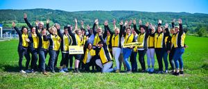 A group of people dressed in yellow and black jackets stand in a field, some raising their hands. In the background, mountains and a clear blue sky are visible. A person in the front holds a yellow sign with the text 'KOMM IN UNSER TEAM!' Another person holds a yellow sign with the hashtag '#team.pflege'.